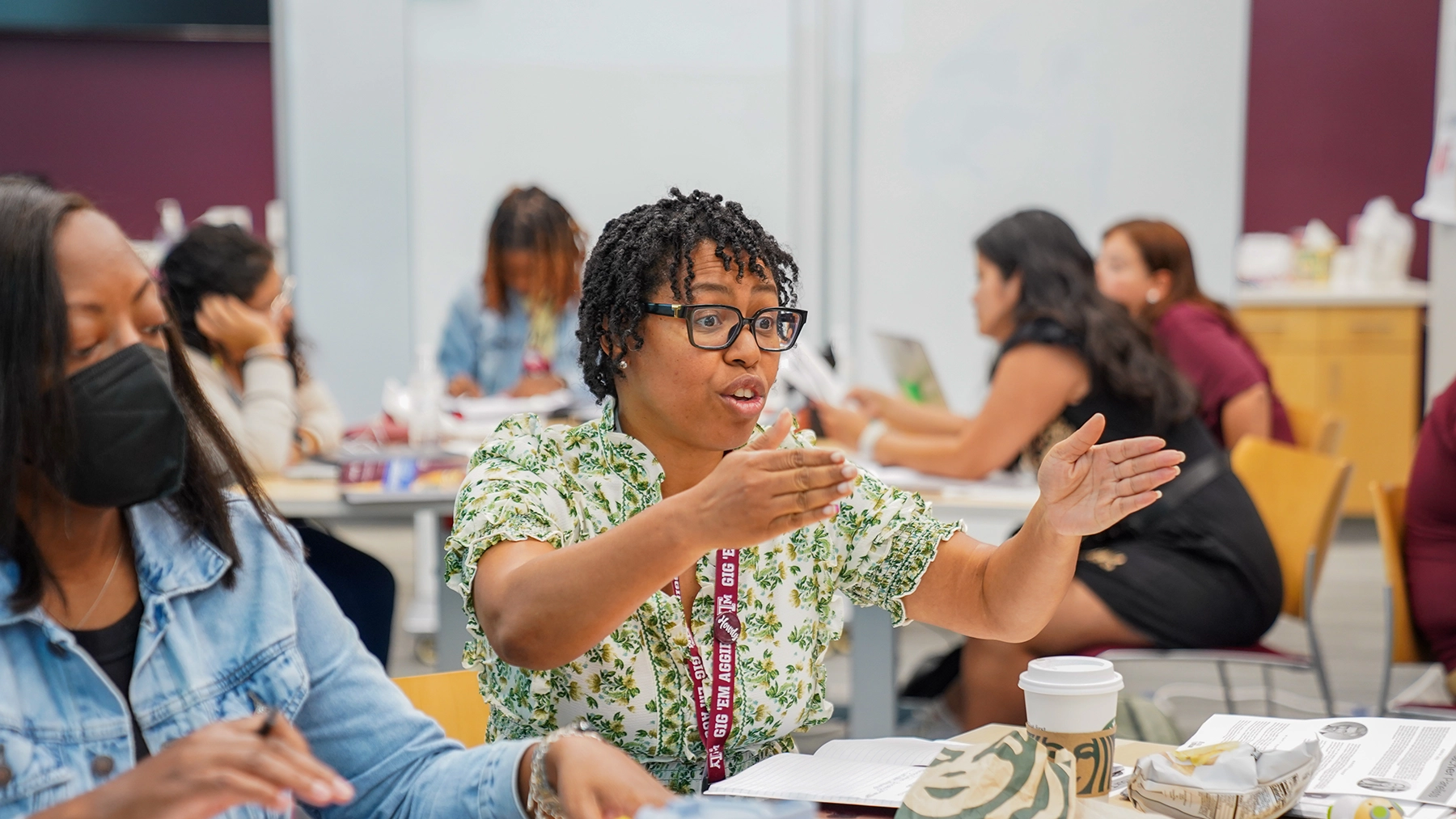 A woman gesturing as she speaks while sitting at a table in a room of people. 
